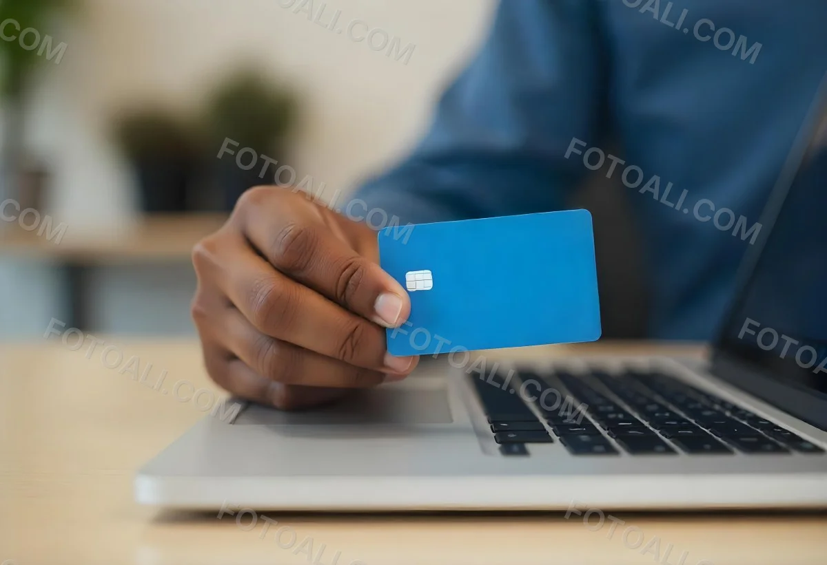 A Brown Skin Hand Holding A Blue Credit Card Over A Laptop The Background Is Blurred And Looks Like An Office 4 Da47