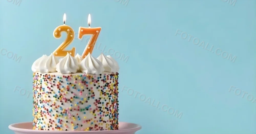 A Cake With White Frosting And Colorful Sprinkles Topped With Two Lit Candles Shaped Like The Numbers 2 And 7 On A White Plate With A Pink Background 2 6670