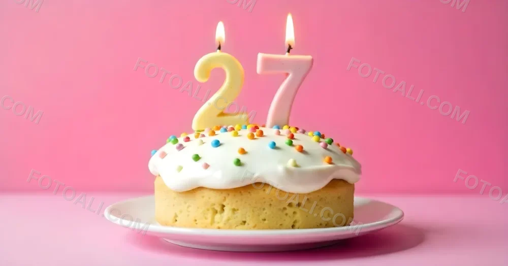 A Cake With White Frosting And Colorful Sprinkles Topped With Two Lit Candles Shaped Like The Numbers 2 And 7 On A White Plate With A Pink Background 22 F21e