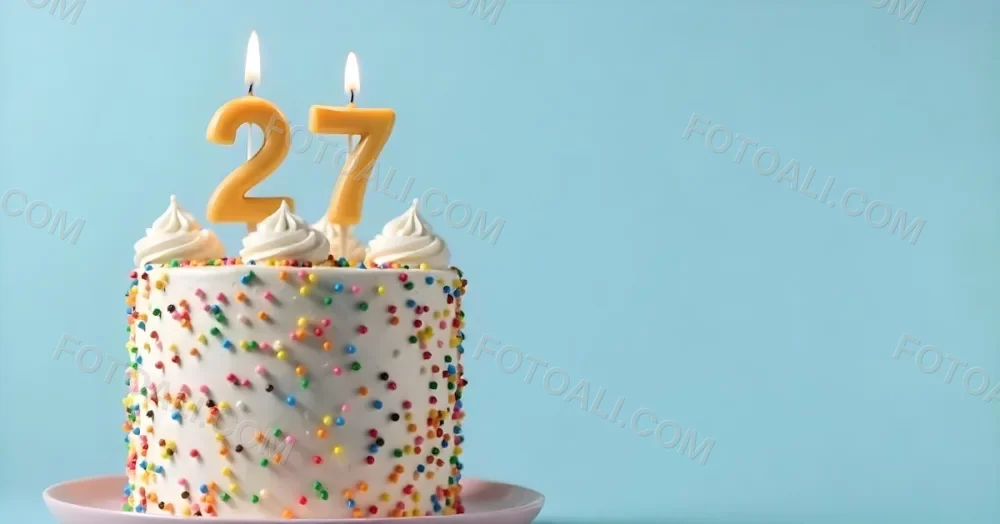 A Cake With White Frosting And Colorful Sprinkles Topped With Two Lit Candles Shaped Like The Numbers 2 And 7 On A White Plate With A Pink Background 7 069e
