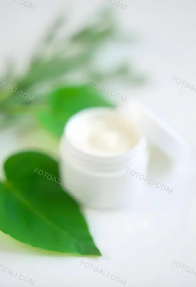 A White Jar With Cream And A Lid Surrounded By Two Green Leaves On A White Surface 91 Dcb8