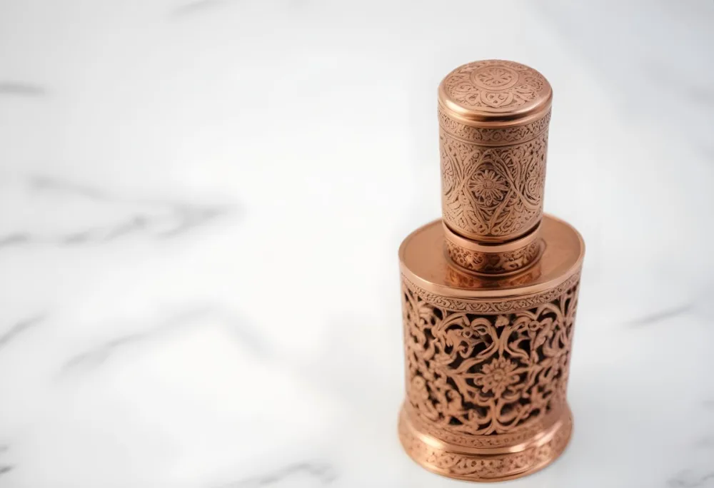 A Decorative Copper Or Bronze Perfume Bottle With An Intricate Floral And Geometric Pattern Engraved On The Surface The Bottle Has A Round Shape With A Narrow Neck And A Decorative Cap On Top Set Against A Marble Counte 33 Fc69