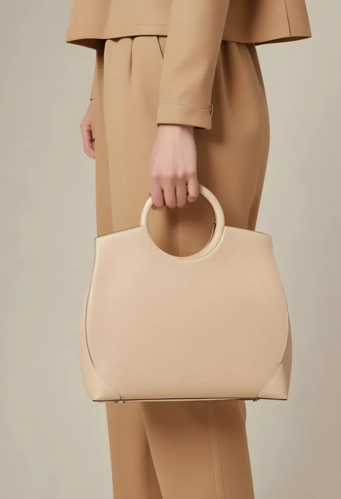 A Hand Holding A Beige Handbag With A Circular Handle The Bag Has A Structured Design And Is Made Of Smooth The Background Is A Neutral Color Complementing The Bag S Tone The Hand Appears To Be Part Of A Person Weari 8 E6c5