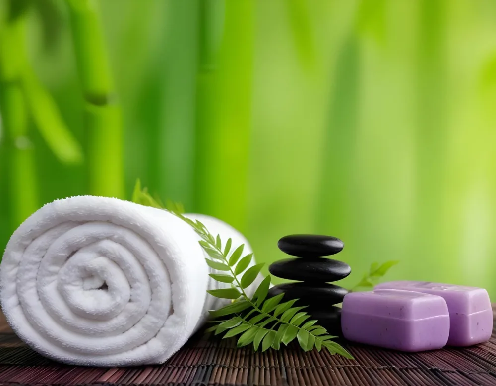 A Rolled White Towel Placed Next To Smooth Black Stones Green Fern Leaves And Small Purple And Pink Soap Bars The Background Features A Blurred Green Bamboo Setting Creating A Tranquil Spa Like Atmosphere 11 Cb2a