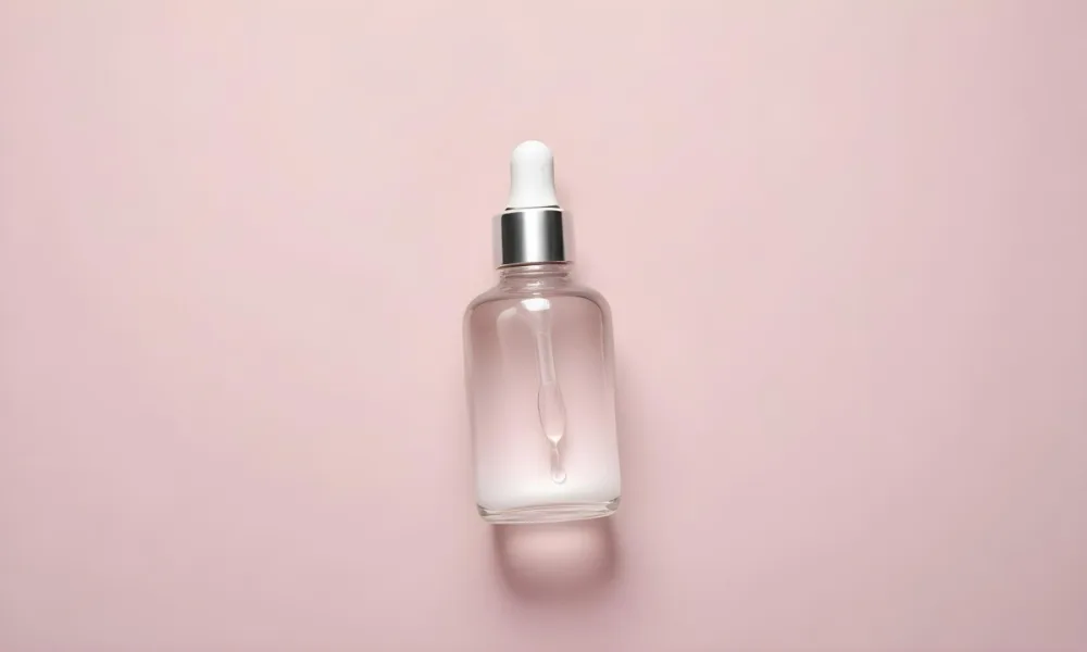 A Transparent Glass Bottle With A White And Silver Dropper Filled With Clear Liquid Laying On A Plain Light Pink Background 11 85aa