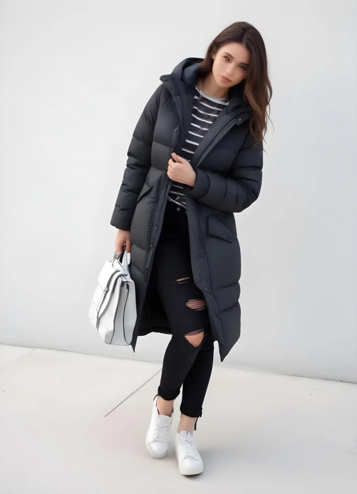 A Young Caucasian Woman With Long Brown Hair Wearing A Dark Gray Hooded Puffer Coat A Striped Shirt And Ripped Black Jeans She Is Holding A White Bag And Appears To Be Standing Outdoors In A Sunny Park 8 65ca