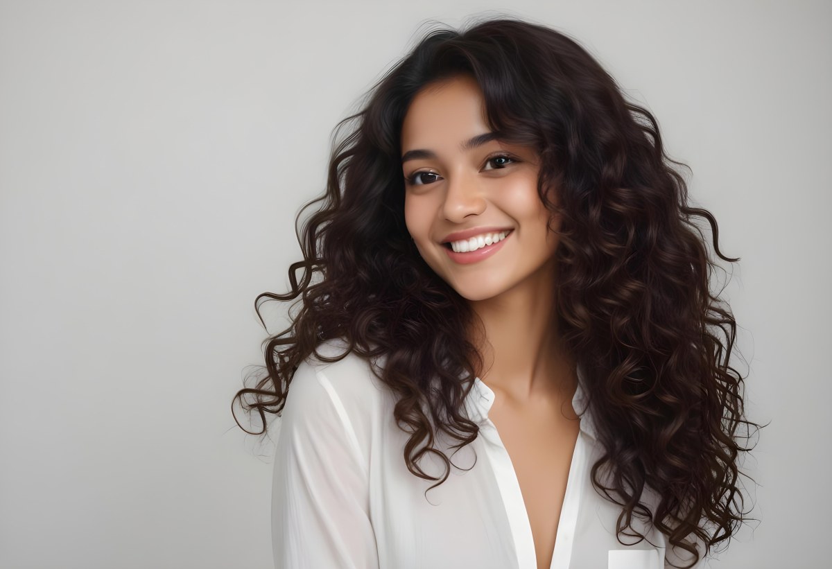 Young Woman Of South Asian Descent With Long Curly Dark Brown Hair Smiling Softly She Is Wearing A White Blouse And Has A Light Background 55 1539