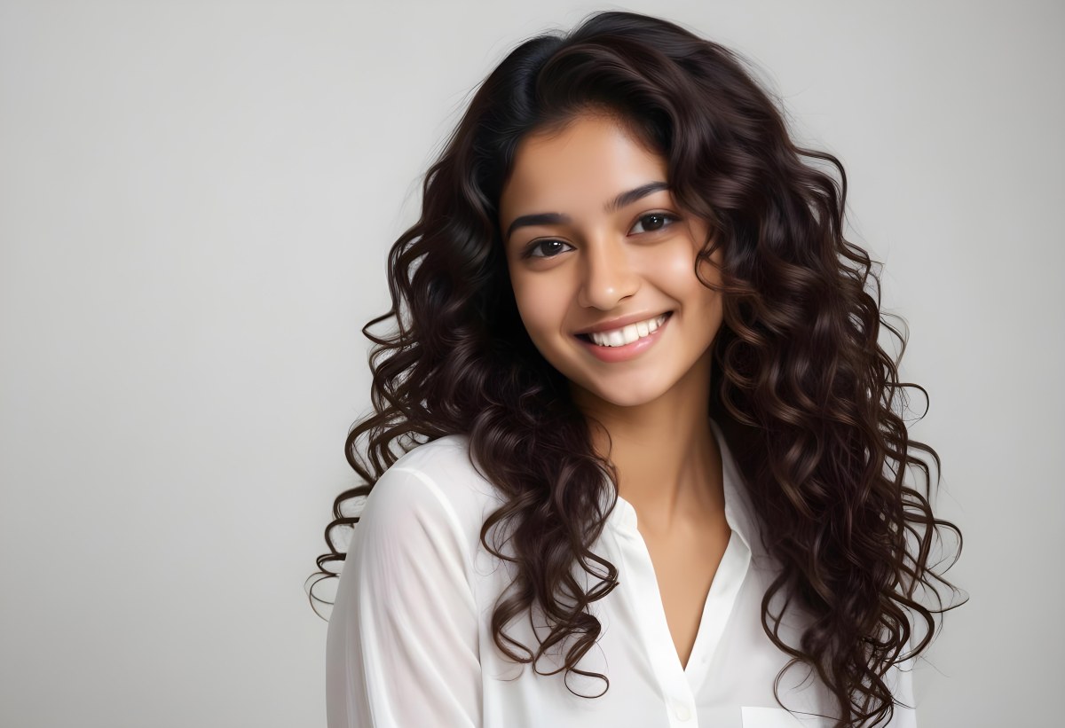 Young Woman Of South Asian Descent With Long Curly Dark Brown Hair Smiling Softly She Is Wearing A White Blouse And Has A Light Background 89 84e5