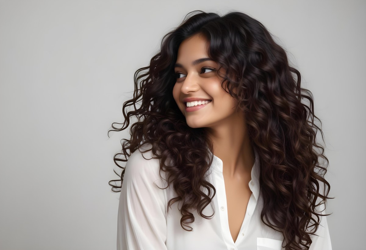 Young Woman Of South Asian Descent With Long Curly Dark Brown Hair Smiling Softly She Is Wearing A White Blouse And Has A Light Background 95 A893