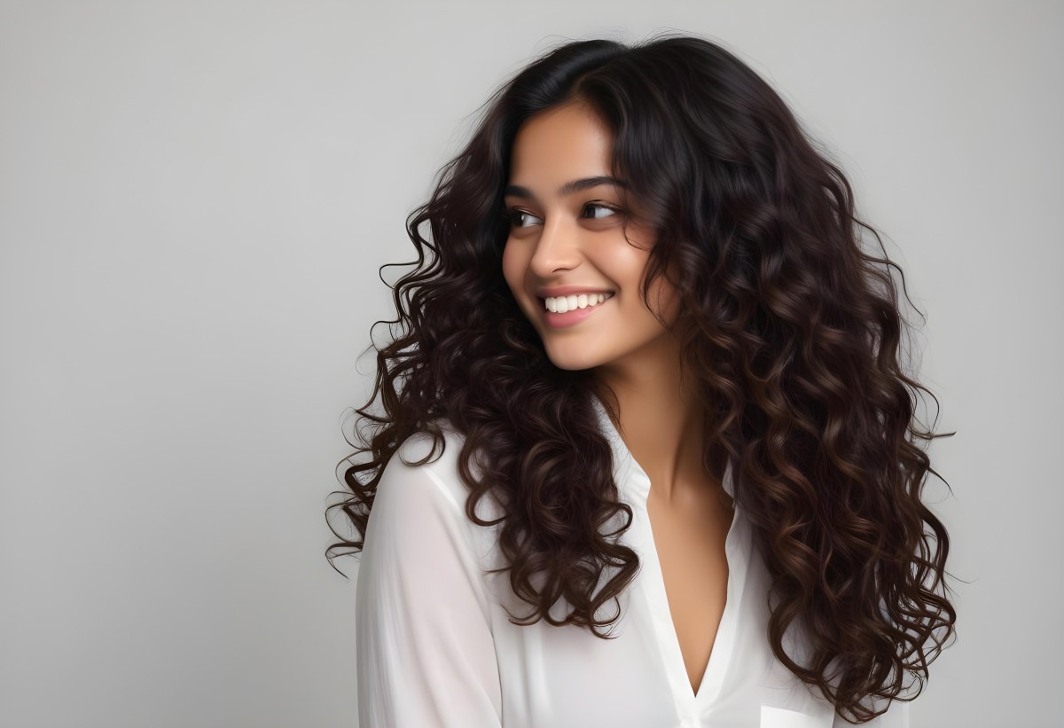 Young Woman Of South Asian Descent With Long Curly Dark Brown Hair Smiling Softly She Is Wearing A White Blouse And Has A Light Background 97 8e7a
