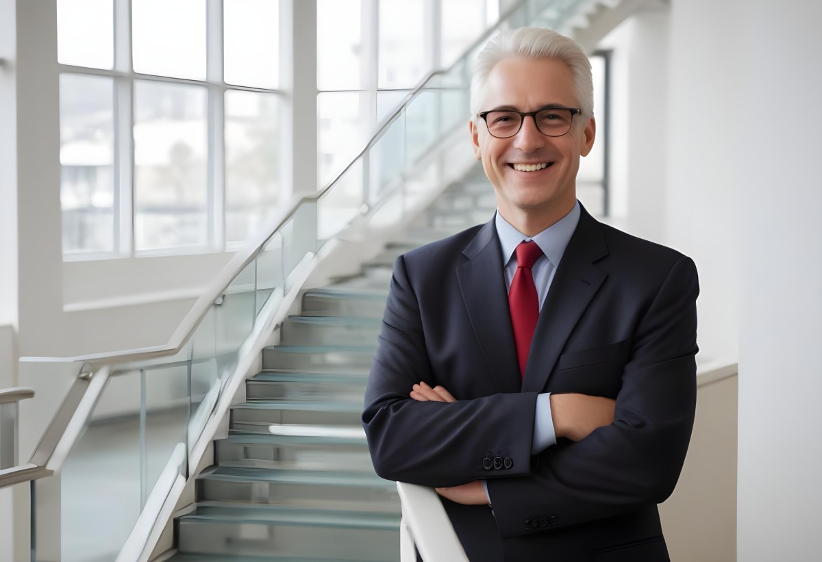 White Man 50 Years Old With White Hair And Glasses Wearing A Suit And A Red Tie Smiling In An Office Environment With A Staircase In The Background 5 015c