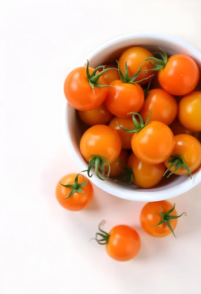 A Bowl Full Of Orange Cherry Tomatoes With Orange Stems Some Tomatoes Scattered On A White Surface Bright Lighting Top Down View 27 Ea87
