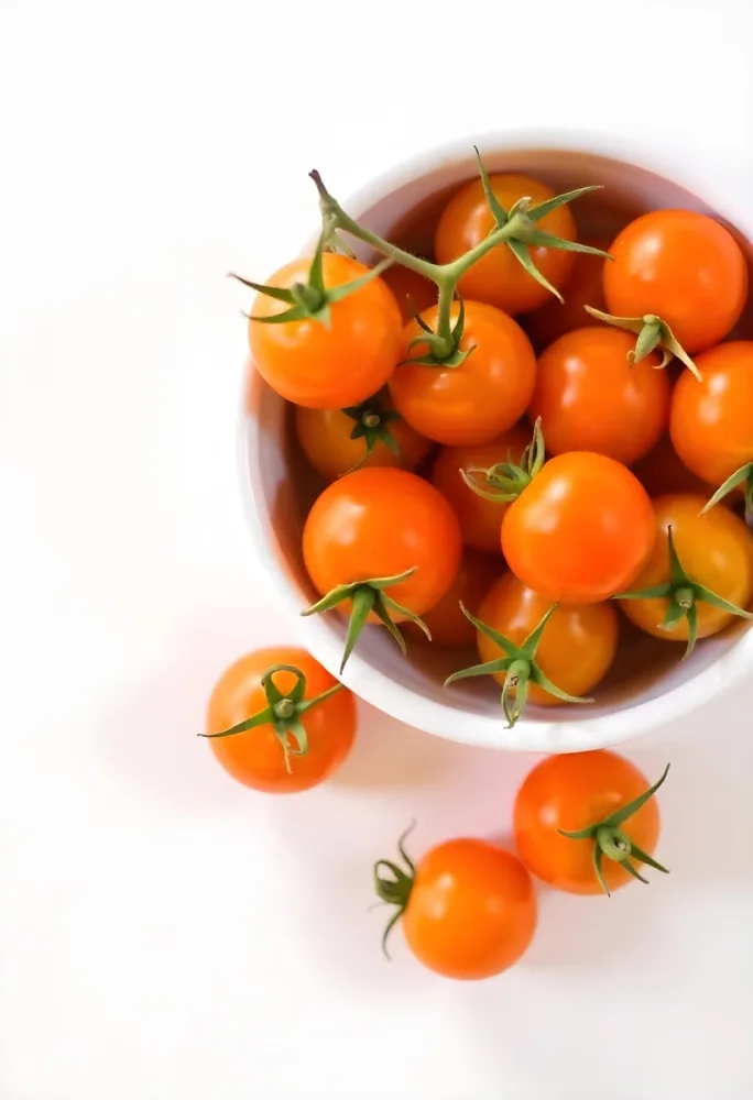 A Bowl Full Of Orange Cherry Tomatoes With Orange Stems Some Tomatoes Scattered On A White Surface Bright Lighting Top Down View 35 E65c