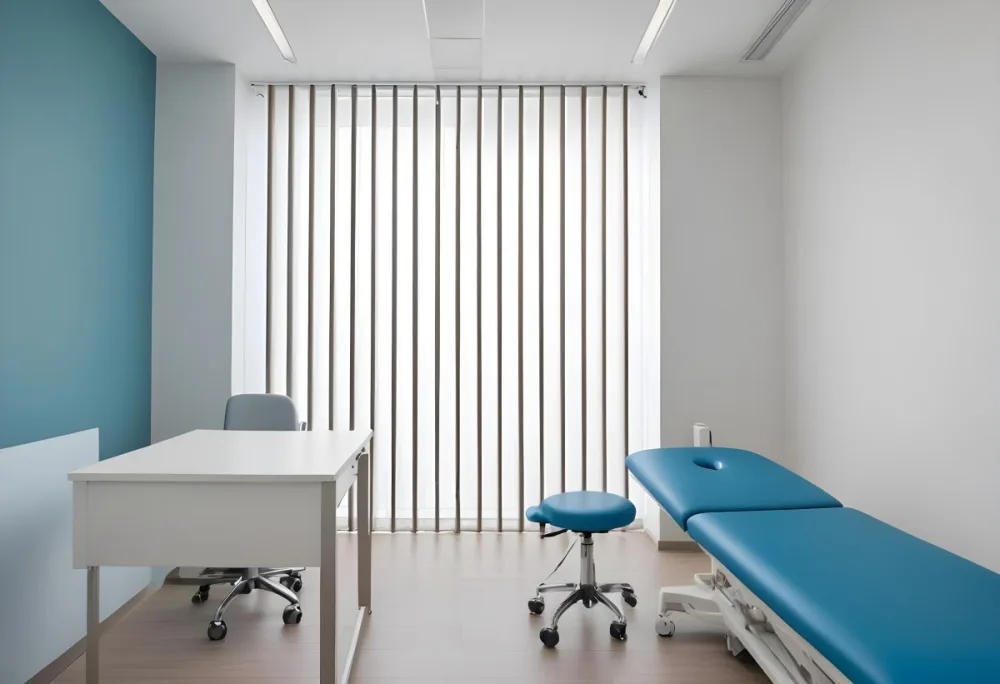 A Modern Medical Examination Room With A White Desk A Blue Examination Table And Vertical Blinds Covering A Window 8 045f