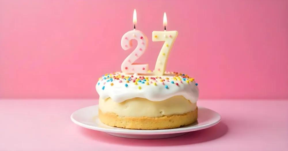 A Cake With White Frosting And Colorful Sprinkles Topped With Two Lit Candles Shaped Like The Numbers 2 And 7 On A White Plate With A Pink Background 12 1fb7