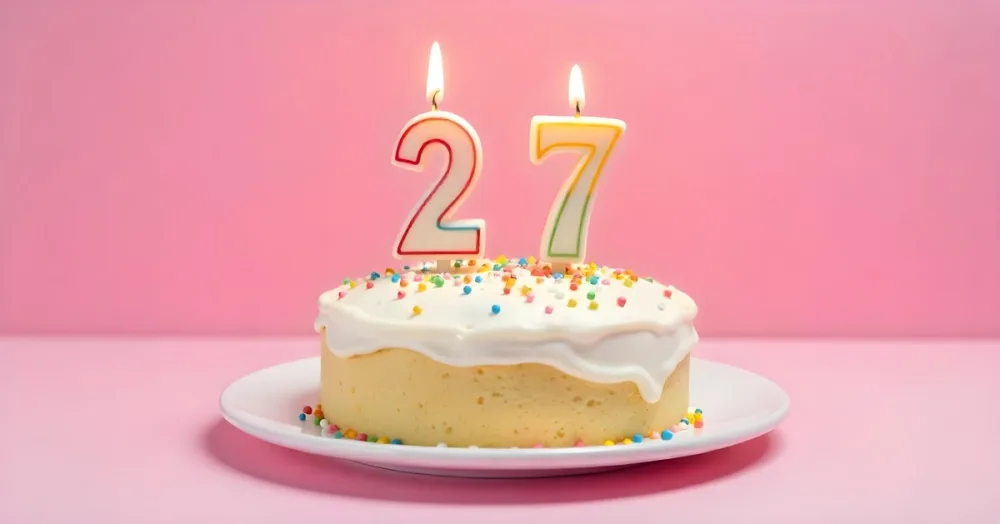 A Cake With White Frosting And Colorful Sprinkles Topped With Two Lit Candles Shaped Like The Numbers 2 And 7 On A White Plate With A Pink Background 13 5b34