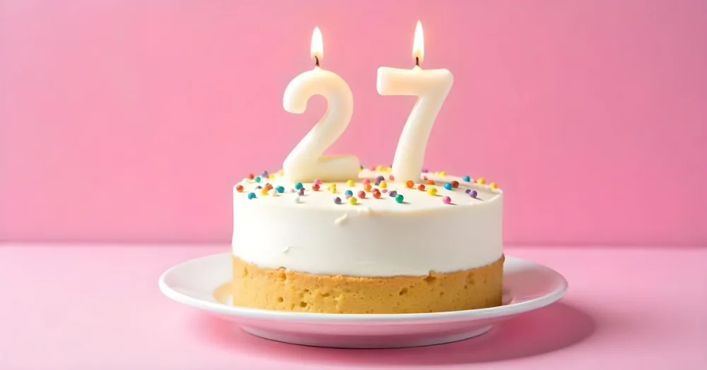 A Cake With White Frosting And Colorful Sprinkles Topped With Two Lit Candles Shaped Like The Numbers 2 And 7 On A White Plate With A Pink Background 16 654a