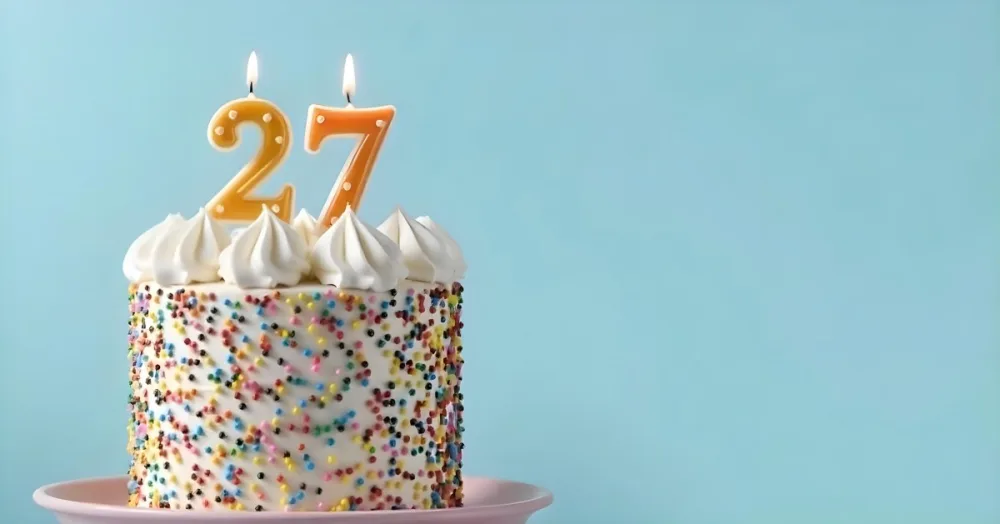 A Cake With White Frosting And Colorful Sprinkles Topped With Two Lit Candles Shaped Like The Numbers 2 And 7 On A White Plate With A Pink Background 2 6670