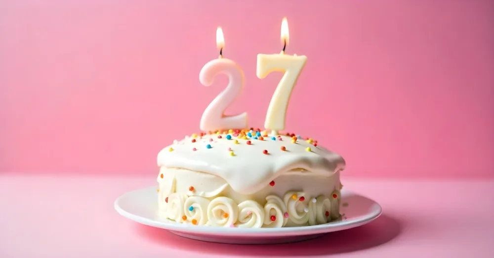 A Cake With White Frosting And Colorful Sprinkles Topped With Two Lit Candles Shaped Like The Numbers 2 And 7 On A White Plate With A Pink Background 20 D792