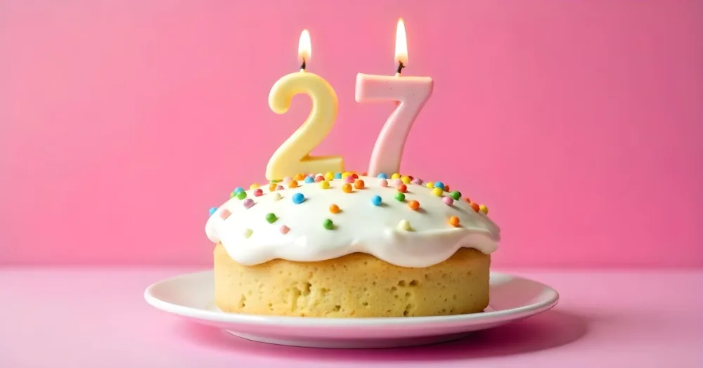 A Cake With White Frosting And Colorful Sprinkles Topped With Two Lit Candles Shaped Like The Numbers 2 And 7 On A White Plate With A Pink Background 22 F21e
