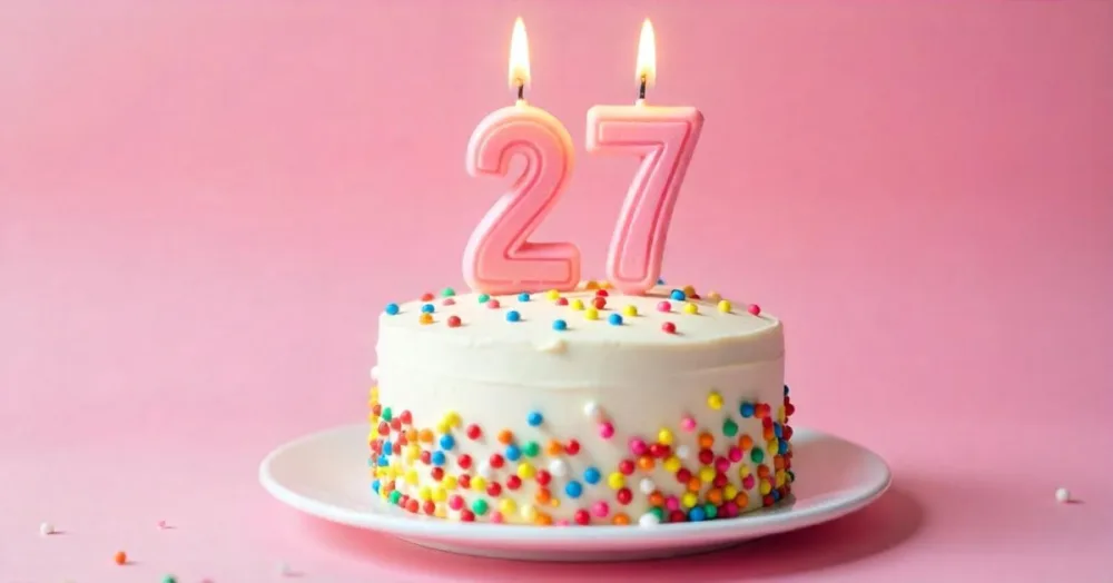 A Cake With White Frosting And Colorful Sprinkles Topped With Two Lit Candles Shaped Like The Numbers 2 And 7 On A White Plate With A Pink Background 23 D1c2