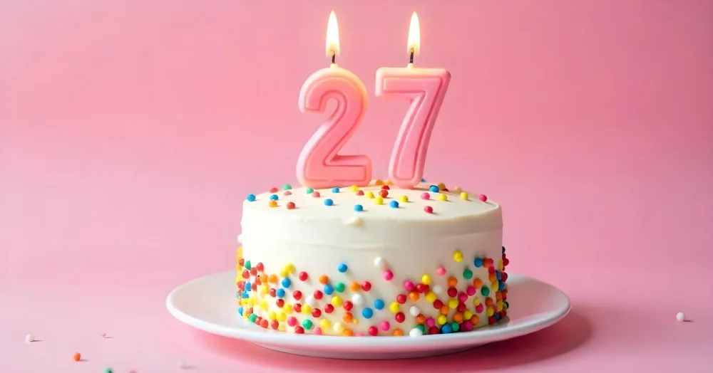 A Cake With White Frosting And Colorful Sprinkles Topped With Two Lit Candles Shaped Like The Numbers 2 And 7 On A White Plate With A Pink Background 24 2531