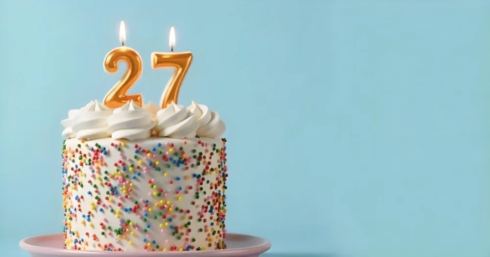 A Cake With White Frosting And Colorful Sprinkles Topped With Two Lit Candles Shaped Like The Numbers 2 And 7 On A White Plate With A Pink Background 3 D111