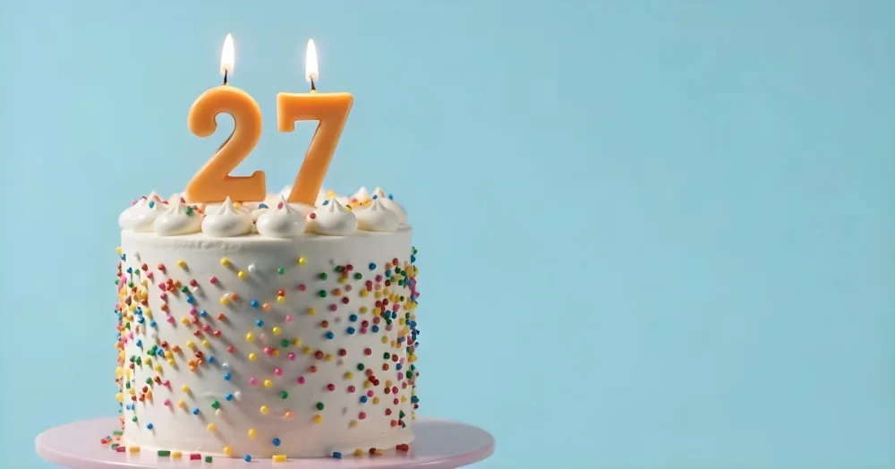 A Cake With White Frosting And Colorful Sprinkles Topped With Two Lit Candles Shaped Like The Numbers 2 And 7 On A White Plate With A Pink Background 4 9334