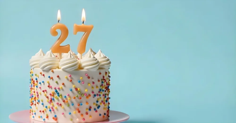 A Cake With White Frosting And Colorful Sprinkles Topped With Two Lit Candles Shaped Like The Numbers 2 And 7 On A White Plate With A Pink Background 5 18db
