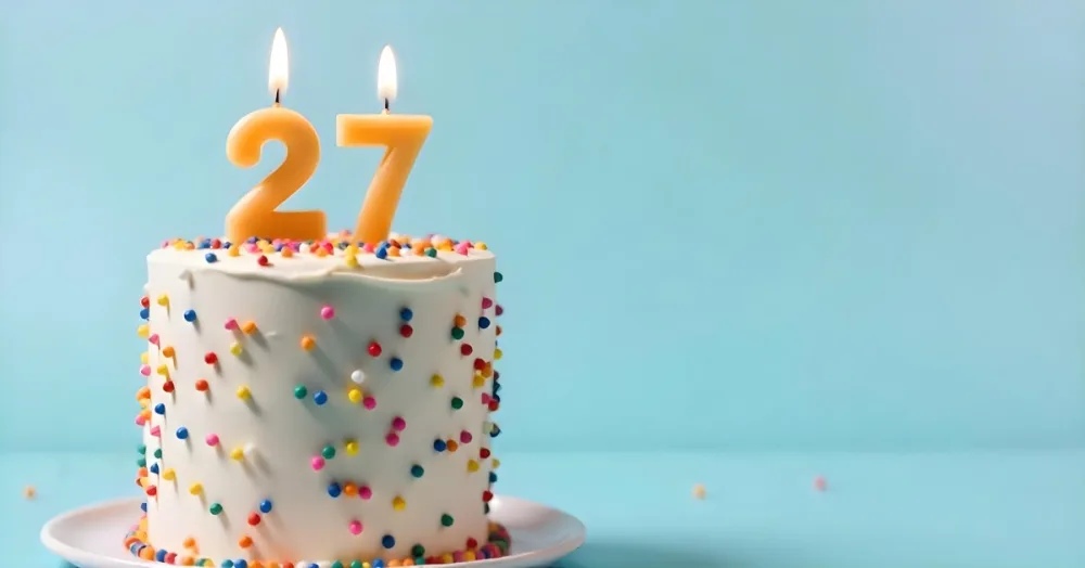 A Cake With White Frosting And Colorful Sprinkles Topped With Two Lit Candles Shaped Like The Numbers 2 And 7 On A White Plate With A Pink Background 6 9cd4