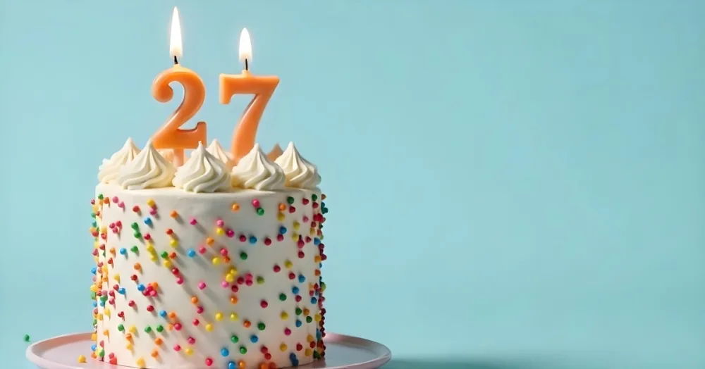 A Cake With White Frosting And Colorful Sprinkles Topped With Two Lit Candles Shaped Like The Numbers 2 And 7 On A White Plate With A Pink Background 8 Ea1c