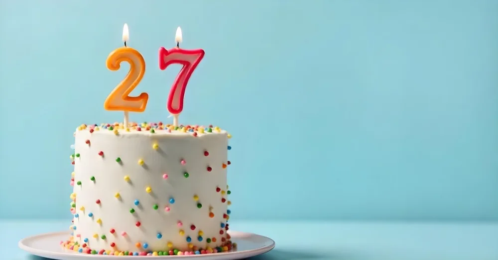 A Cake With White Frosting And Colorful Sprinkles Topped With Two Lit Candles Shaped Like The Numbers 2 And 7 On A White Plate With A Pink Background 9 2f34