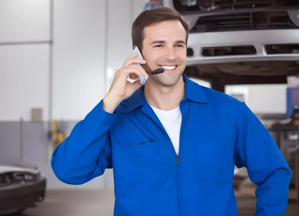A Caucasian Male Approximately 30 Years Old With Short Brown Hair Wearing A Blue Work Uniform He Is Smiling And Talking On A Smartphone The Background Features A Garage Setting With Cars And Tools Visible 3 48c6