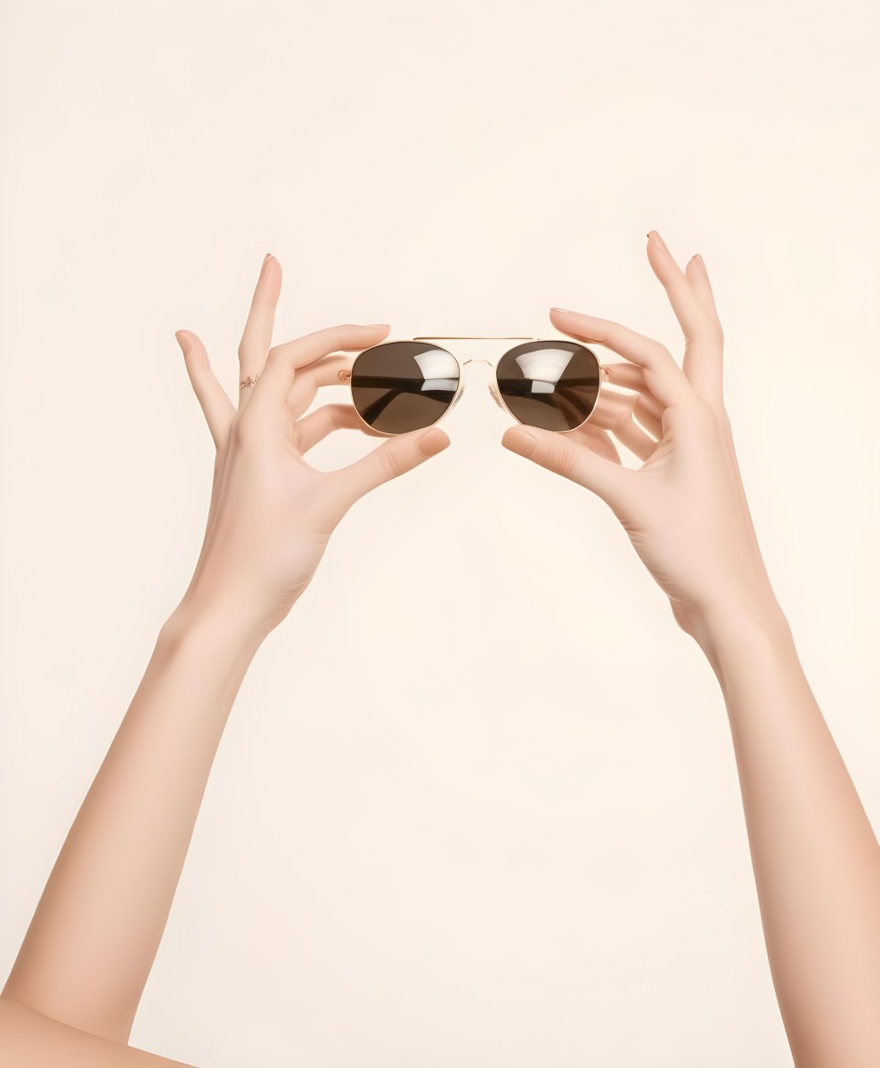 A Woman S Hands Holding A Pair Of Gold Framed Sunglasses Against A Plain White Background 5 05a6