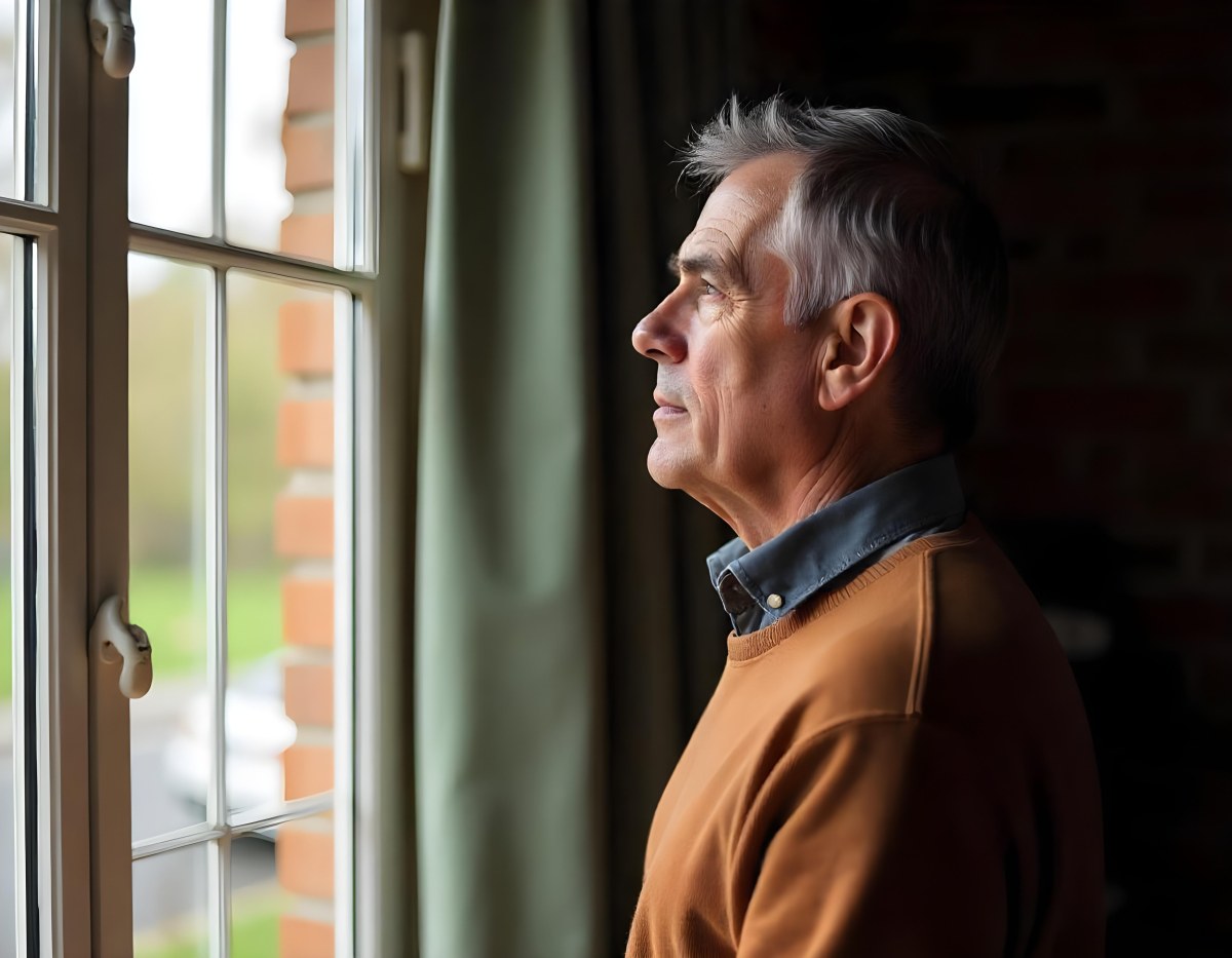 A Middle Aged Caucasian Man With Short Gray Hair Standing By A Window In A Bedroom Looking Outside At A Cityscape During Daytime 2 032e