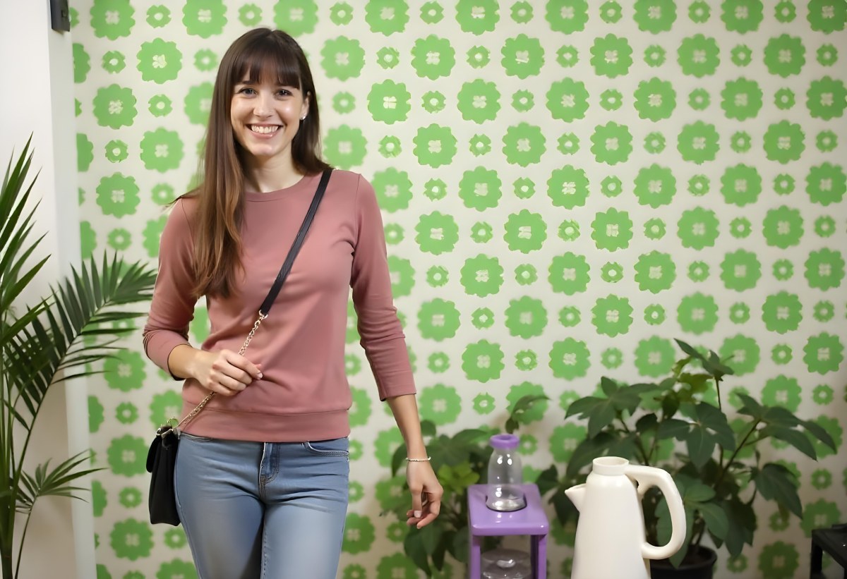 A Young Caucasian Woman With Long Brown Wavy Hair Wearing A Pink Sweater Smiling In Front Of A Green Background With White Flowers 8 F794