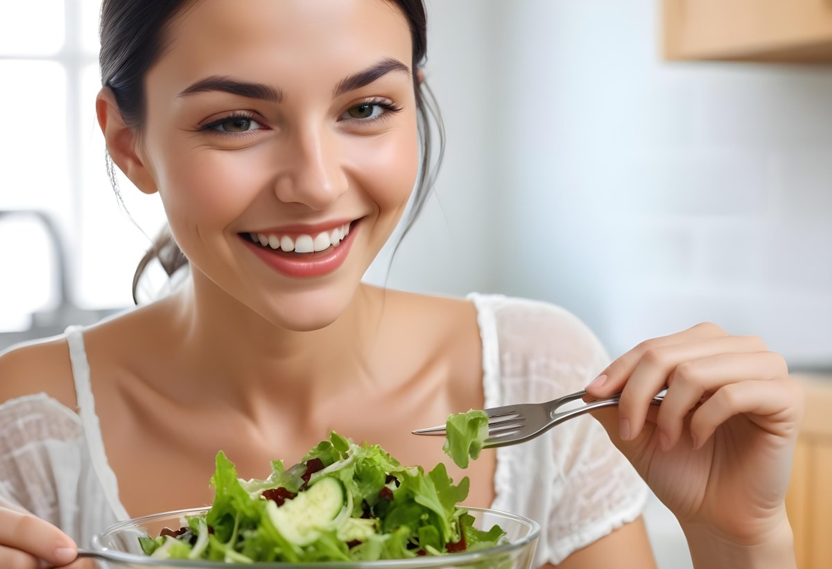 A Young Caucasian Woman With Long Dark Hair Eating A Fresh Green Salad In A Brightly Lit Kitchen Smiling And Looking Down At The Bowl 2 190a