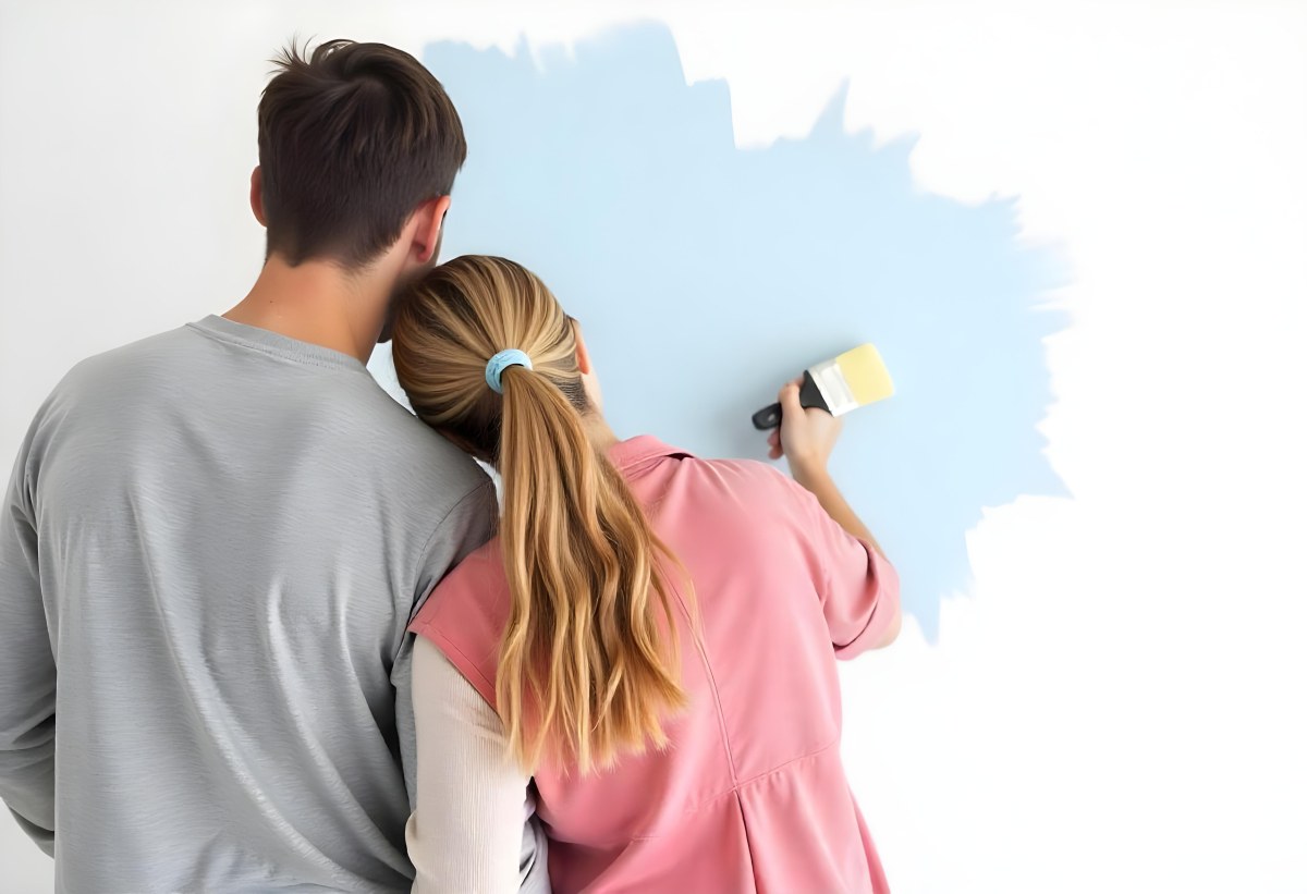 A Young Couple Painting A Wall Together In Their Home The Man Holding A Paint Roller And The Woman Standing Close Behind Her Daytime With Sunlight Streaming In In The Background 6 88fb