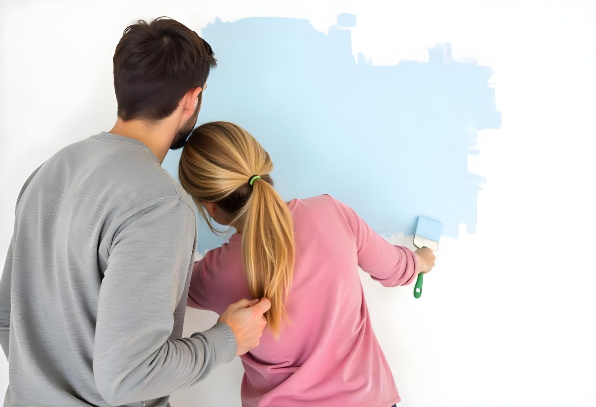 A Young Couple Painting A Wall Together In Their Home The Man Holding A Paint Roller And The Woman Standing Close Behind Her Daytime With Sunlight Streaming In In The Background 7 E5f1