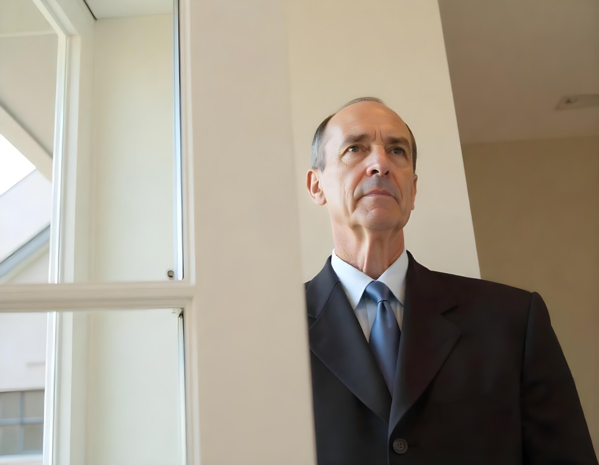 An Elderly Caucasian Man With Gray Hair Wearing A Dark Suit And Tie Standing In Front Of A Window 1 D875
