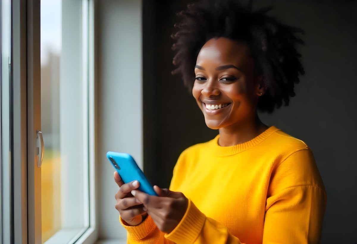 A Black Woman With Dark Skin Around 30 Years Old Wearing A Yellow Sweater And Holding A Blue Smartphone In Front Of A Window 11 7c6b
