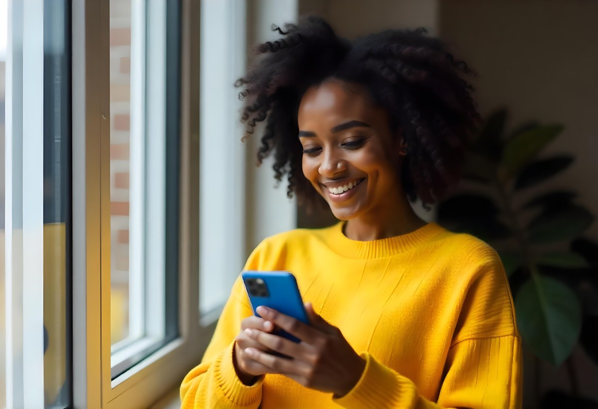 A Black Woman With Dark Skin Around 30 Years Old Wearing A Yellow Sweater And Holding A Blue Smartphone In Front Of A Window 15 F9f3
