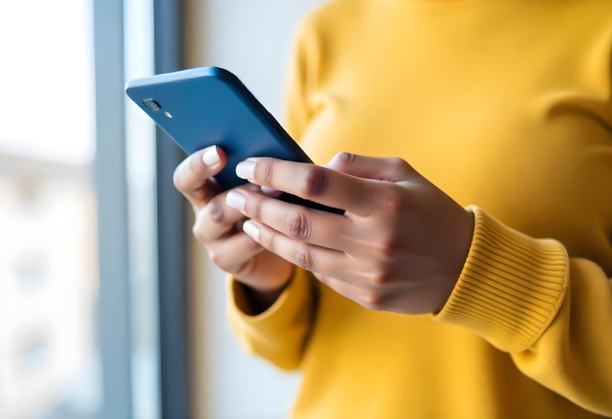 A Black Woman With Dark Skin Around 30 Years Old Wearing A Yellow Sweater And Holding A Blue Smartphone In Front Of A Window 16 7e4e