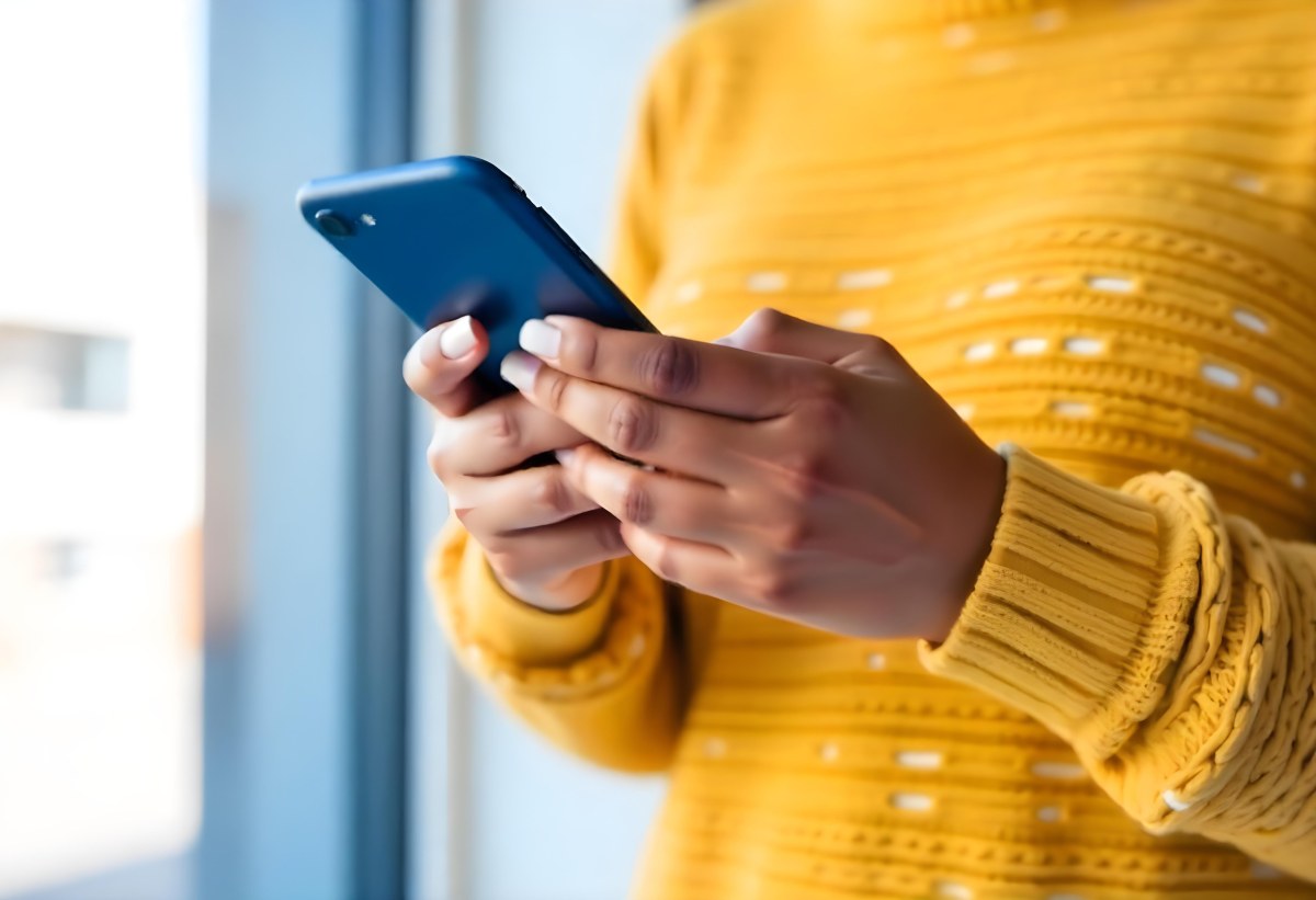 A Black Woman With Dark Skin Around 30 Years Old Wearing A Yellow Sweater And Holding A Blue Smartphone In Front Of A Window 2 63af