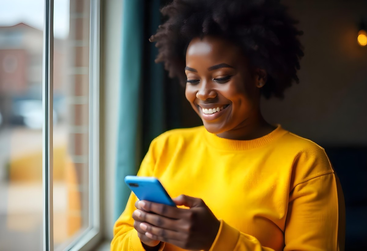 A Black Woman With Dark Skin Around 30 Years Old Wearing A Yellow Sweater And Holding A Blue Smartphone In Front Of A Window 20 B928