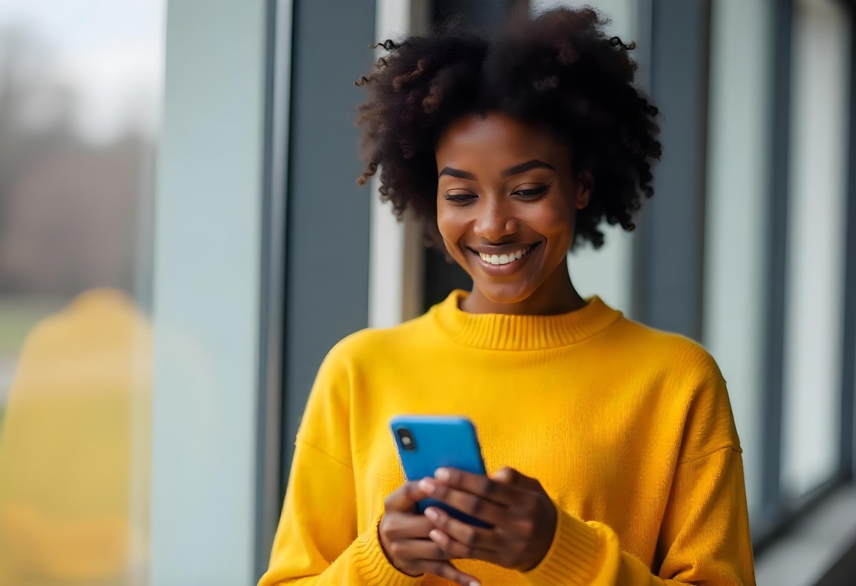 A Black Woman With Dark Skin Around 30 Years Old Wearing A Yellow Sweater And Holding A Blue Smartphone In Front Of A Window 21 7afa