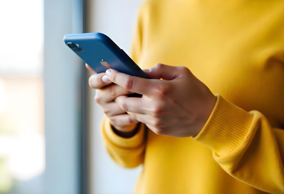 A Black Woman With Dark Skin Around 30 Years Old Wearing A Yellow Sweater And Holding A Blue Smartphone In Front Of A Window 22 F924