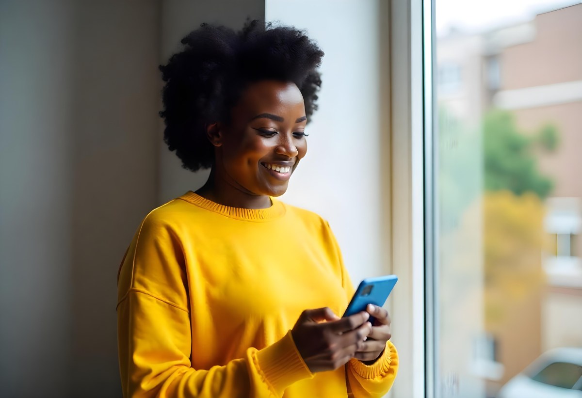 A Black Woman With Dark Skin Around 30 Years Old Wearing A Yellow Sweater And Holding A Blue Smartphone In Front Of A Window 23 D27b