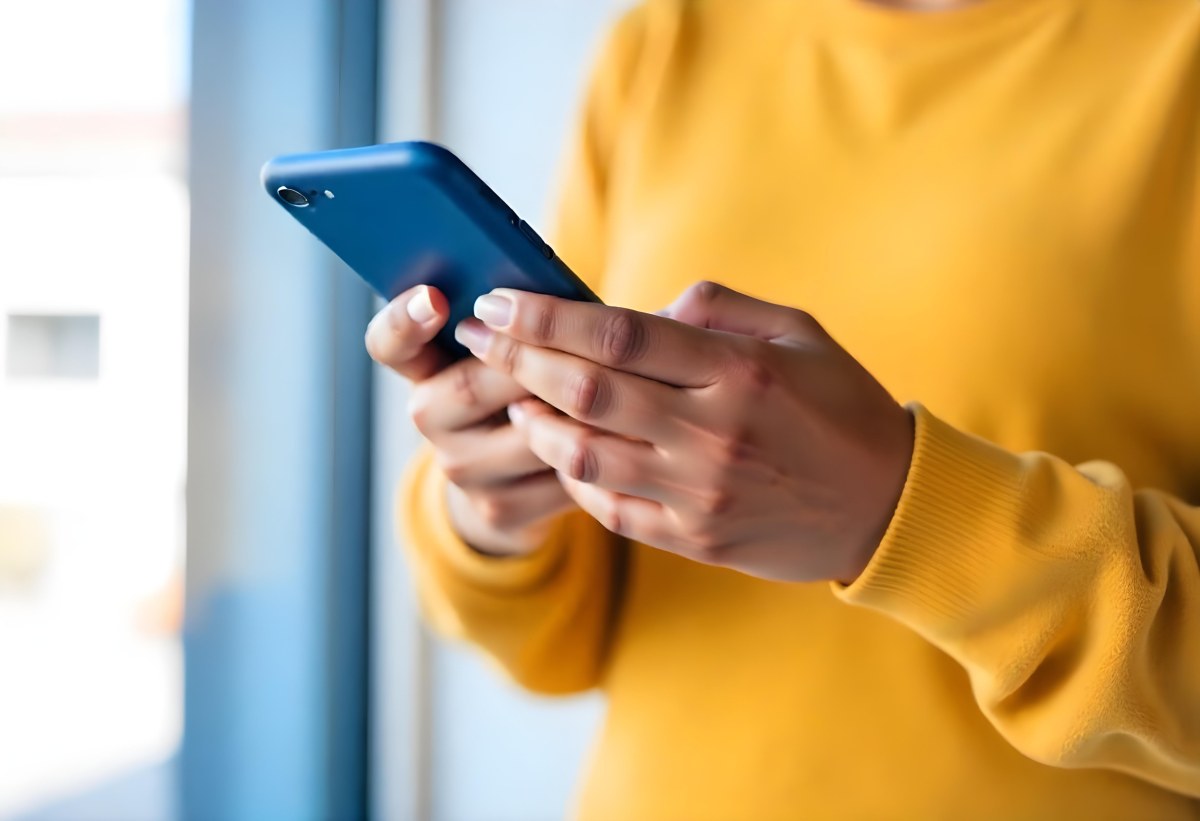 A Black Woman With Dark Skin Around 30 Years Old Wearing A Yellow Sweater And Holding A Blue Smartphone In Front Of A Window 3 Ce09