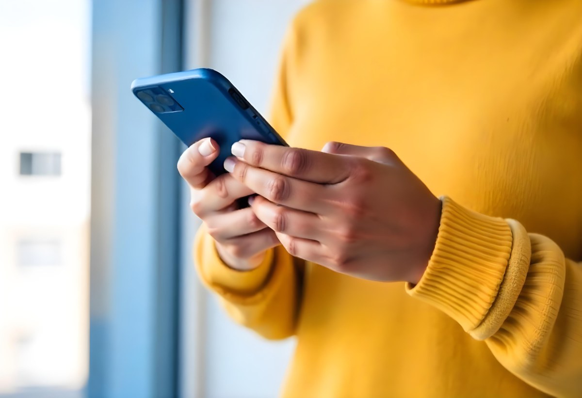 A Black Woman With Dark Skin Around 30 Years Old Wearing A Yellow Sweater And Holding A Blue Smartphone In Front Of A Window 4 67db